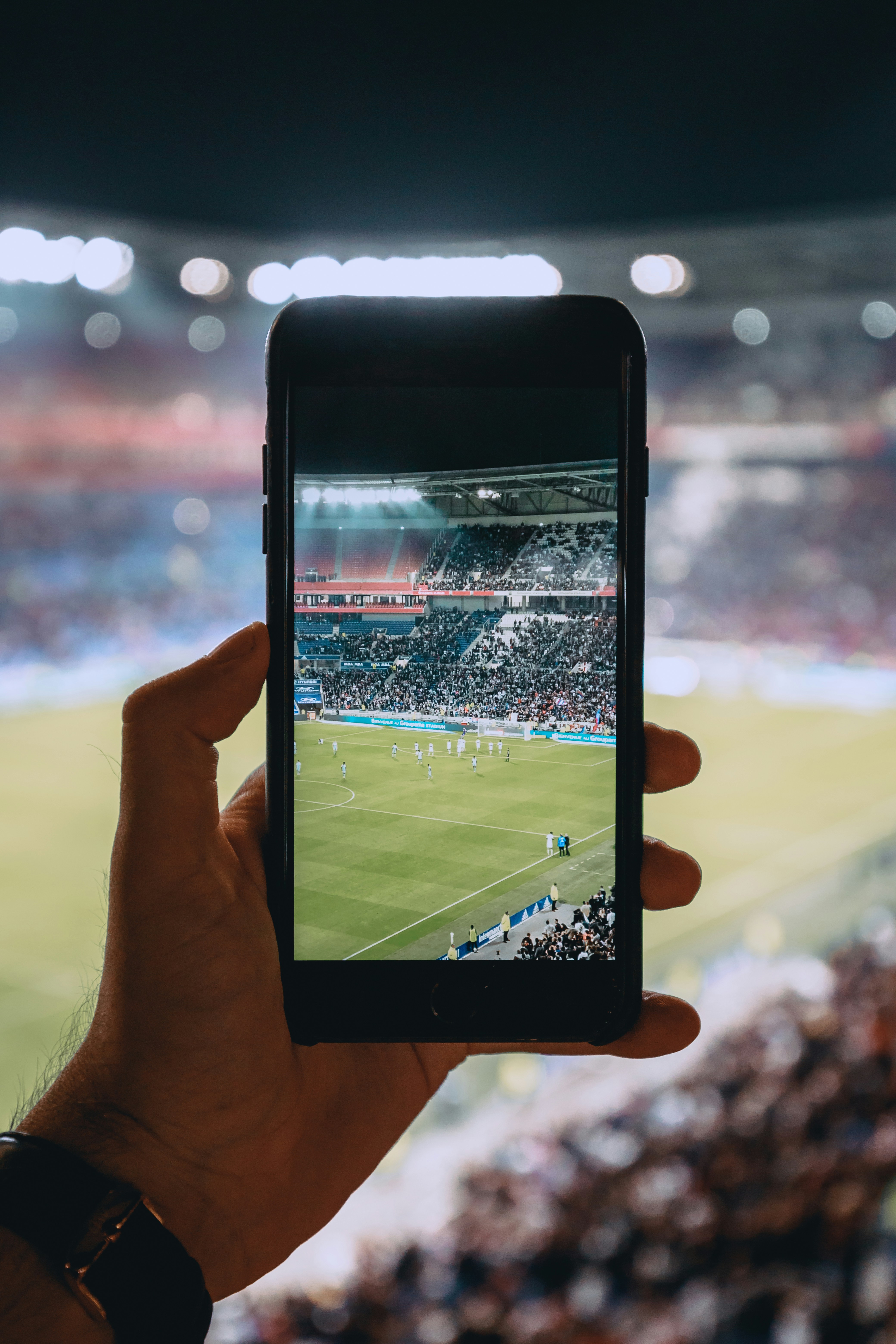 Image shows a hand holding a smartphone, which has the camera app open capturing a football match at a stadium. 